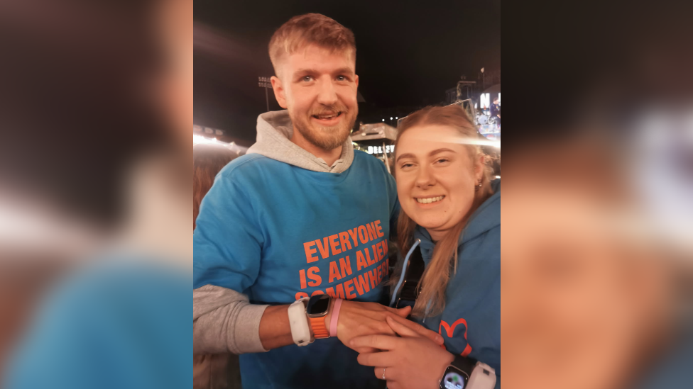 A man and a woman - Nick Bartrupe, and Amber Hird - stood in an embrace holding hands. Nick has short brown hair and is wearing a blue t-shirt with orange writing over a grey hoodie. He has a watch and a white wristband on his hand. Amber has long light brown hair and is wearing a blue hoodie. She has a ring on her finger. 