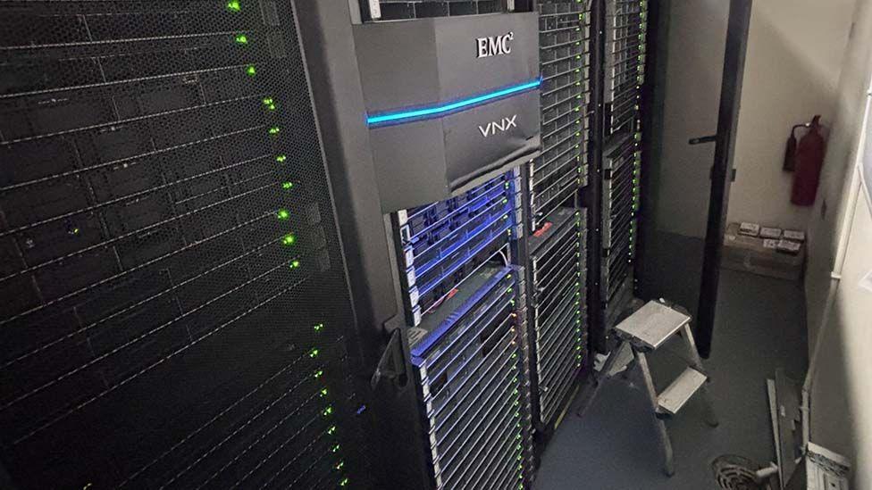 A row of computer servers, tall stacked black computer cabinets with multiple trays and flashing green lights