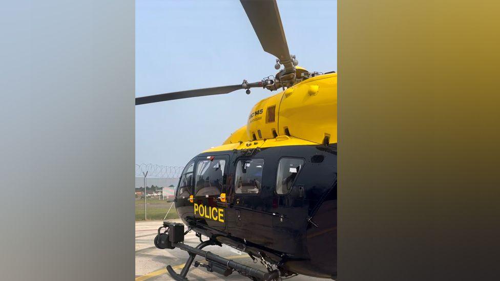 A police helicopter on the ground. The photo only shows the front of the vehicle and two rotas, not the tail. The lower half is black and the upper half bright yellow, with POLICE in yellow on the lower half. You can see two rotas, and in the distance is a high barbed wire fence.