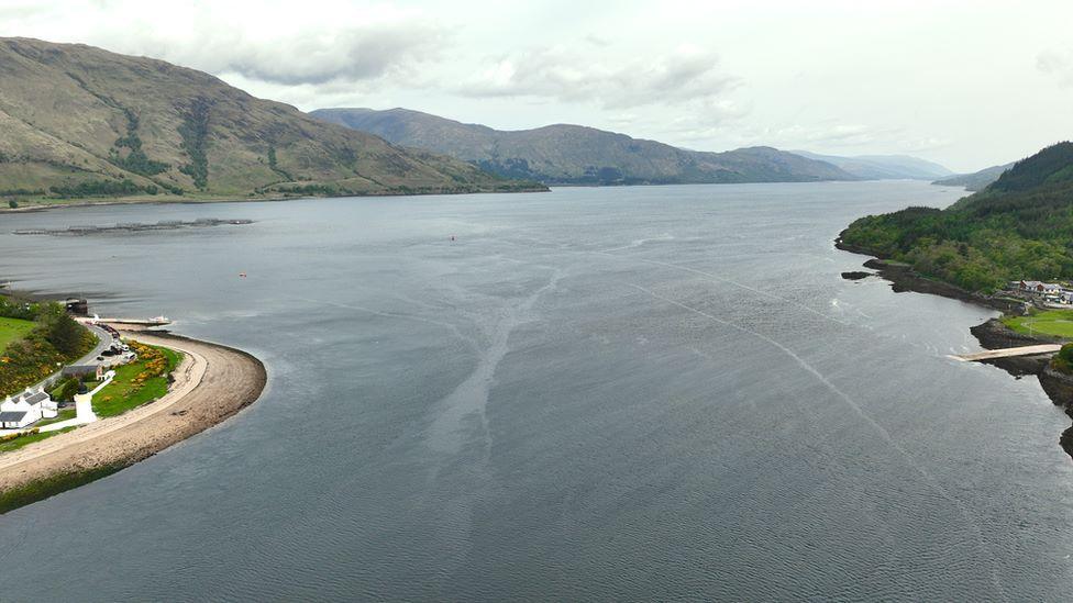 An aerial view of the Corran Narrows on Loch Linnhe. The sea looks grey between two points of land. There is the wider loch, and hills, in the distance.