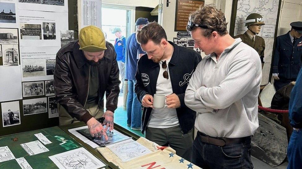 Three men, looking at paperwork inside the museum. They are photos and pictures on all the walls, and dressed mannequins, wearing uniforms. The man on the left has tattoos on his hands, is wearing a green cap, a black leather jacket, the man in the middle has glasses round his white T-shirt, a dark jacket and the man on the right has a grey top on and dark trousers. 