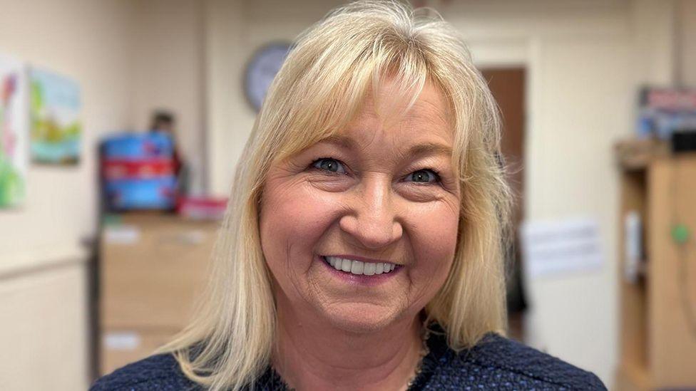 A woman with blonde shoulder-length hair wearing a blue top, standing in an office with filing cabinets in the background. She is smiling at the camera.