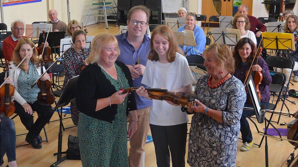 Natasha being handed the violin by Trevor's daughters