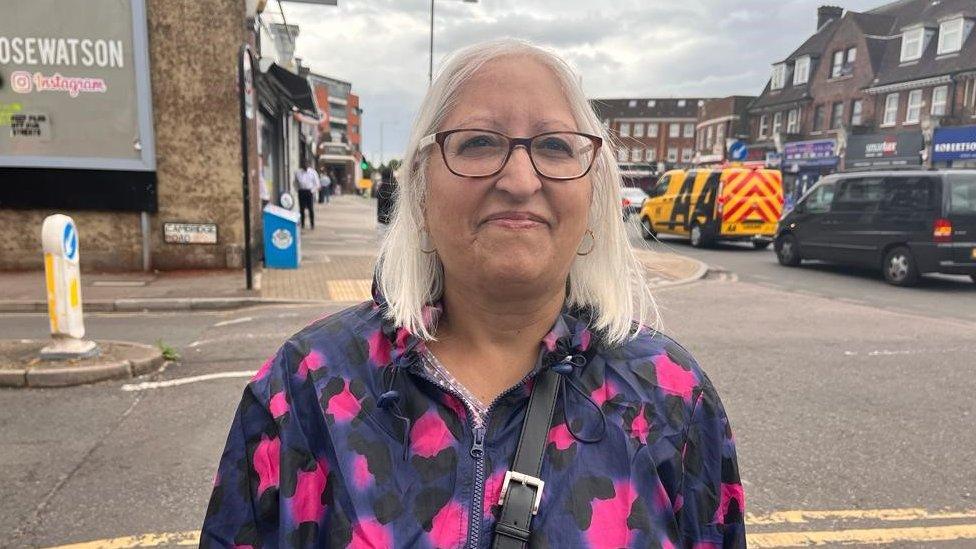 Pushpa, a woman with grey hair, glasses and a blue and pink jacket.