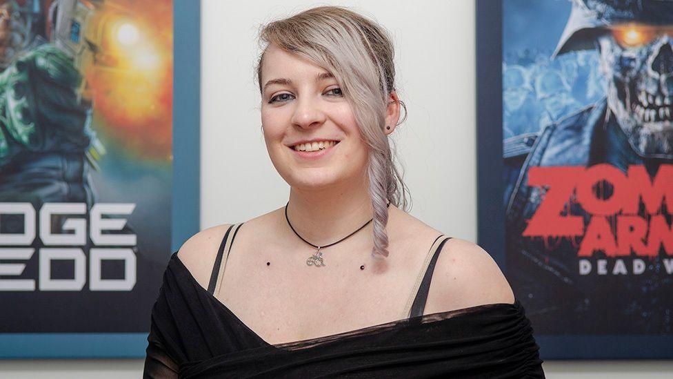 Cari, wearing a black top, smiling, with two posters of video games in the background in blue frames.