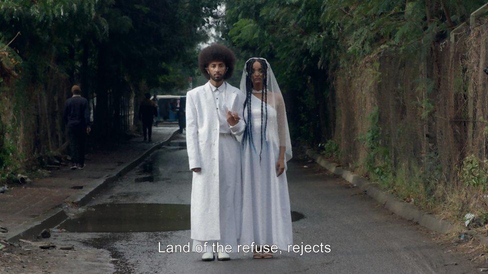 A still from "Except this time nothing returns from the ashes", showing two characters dressed in wedding attire. They are holding hands and looking directly into the camera. The caption reads "land of the refuse, rejects"