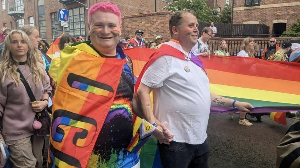 People taking part in the parade, wearing outfits filled with different colours and patterns. One person has pink hair, holding hands with another person next to them.