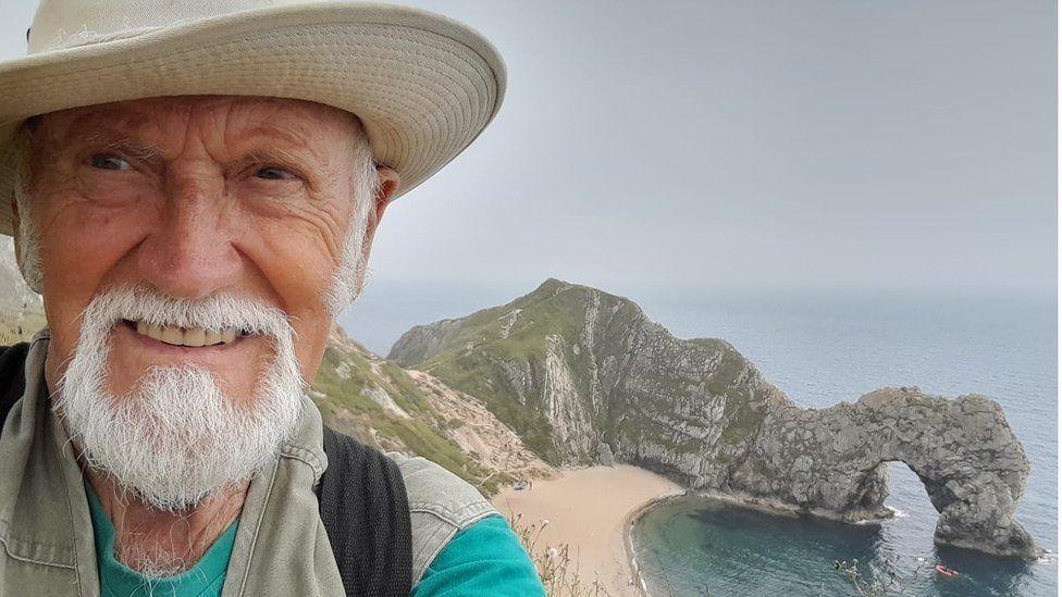 Derek Adams with short white hair and beard, wearing a light-coloured hat with a wide brim. He is looking at the camera with a coastline behind him, showing green and grey rocks and a sand path. There is a natural stone arch above the water to the right.