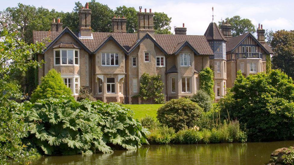 York Cottage on the Sandringham estate. It is a large, brown-brick two-floor property set near a lake. It has a brown roof and a turret on the right-hand side.