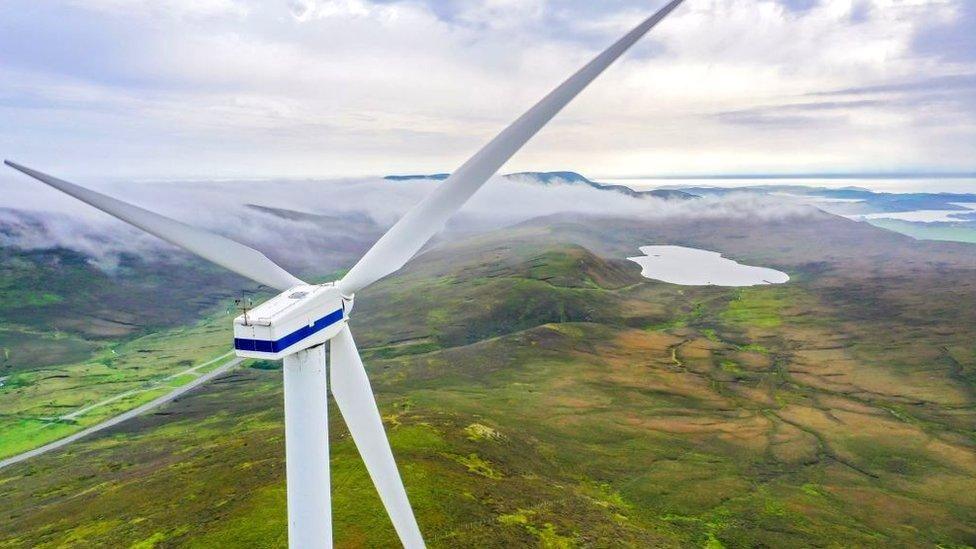 A wind turbine in the foreground looks out over a picturesque Scottish glen, complete with a loch and low clouds over mountaintops