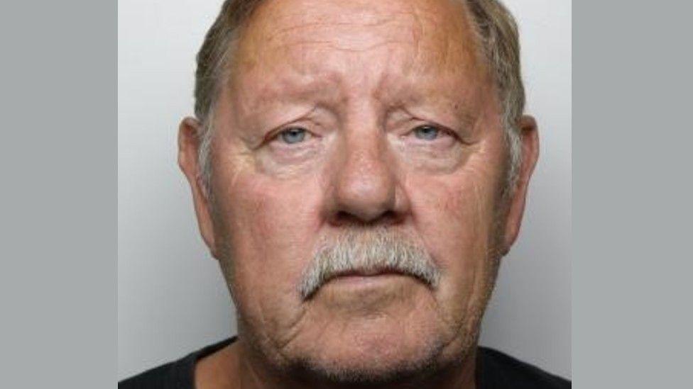 Police custody picture of a man with a grey moustache and hair. He is looking directly at the camera and is wearing a black T-shirt.