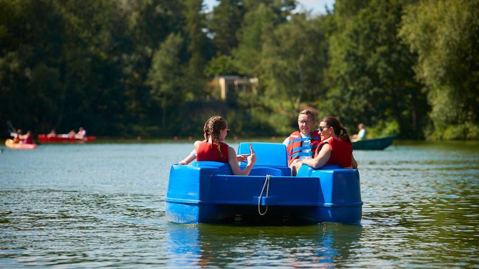 A family of three in a pedalo enjoy the sunshine on a small loch with trees in the background along the shoreline