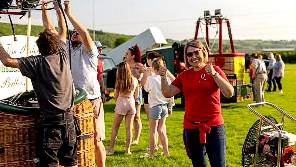Laura Davies stands to the right of the picture. She has bobbed light brown, blonde streaked hair and wears a red short-sleeved T-shirt with a small balloon logo on the left side, navy jeans and a red jumper tied around her waist. She is standing on a grassy field. To her left two men in shorts and t-shirts are adjusting fastenings above a wicker balloon basket. To her right is a fan used to inflate balloons. In the background there is another balloon basket standing on a low trailer. About 10 people can be seen, many in shorts. It is a sunny day.