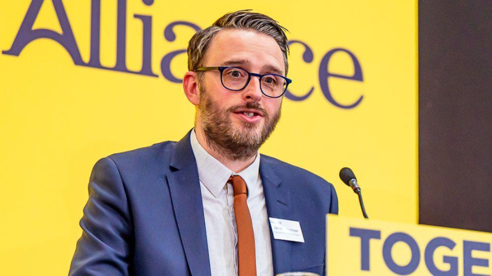 Nick Matheson has a grey beard, grey/whitish hair that is gelled into a spike and grey round glasses. He is wearing a navy shirt, white shirt and orange tie, and behind him is the yellow white Alliance banner