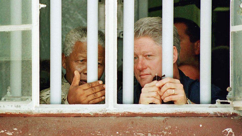 Bill Clinton (R) and Nelson Mandela peer out through the bars of a cell on Robben Island in 1998