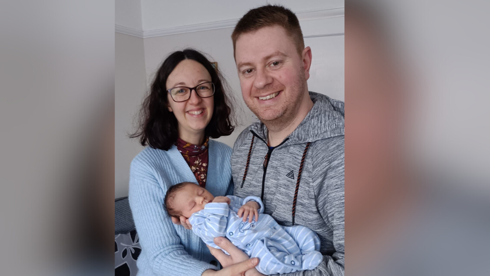 Brunette woman, wearing glasses and pale blue cardigan stands with sandy haired man wearing a grey hoodie. Both are smiling and he is holding a sleeping newborn baby in a pale blue babygrow.