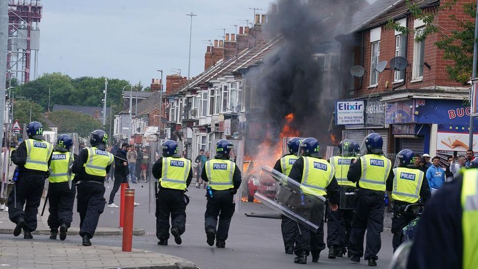 Police wearing protective riot gear approach protesters in Middlesbrough as a car parked on the street is ablaze