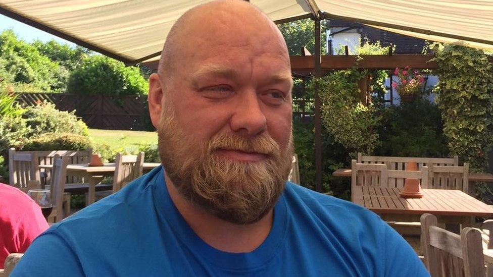 A bald man with a large beard and wearing a blue shirt sits in an outdoor seating area on a sunny day. Wooden tables and chairs can be seen behind him, as well as a garden.