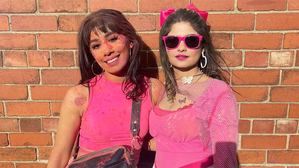 Shaz and Mahum - two female friends standing next to each other. Shaz on the left is smilng and has pink powdered colour on her face, with silver hoop earrings, wearing a pink vest top with a black bag strap hanging. Mahum on the right is wearing sunglasses with pink rims, white hoop earrings and a pink top, with a pink bow in her hair. The background is of a brick wall.