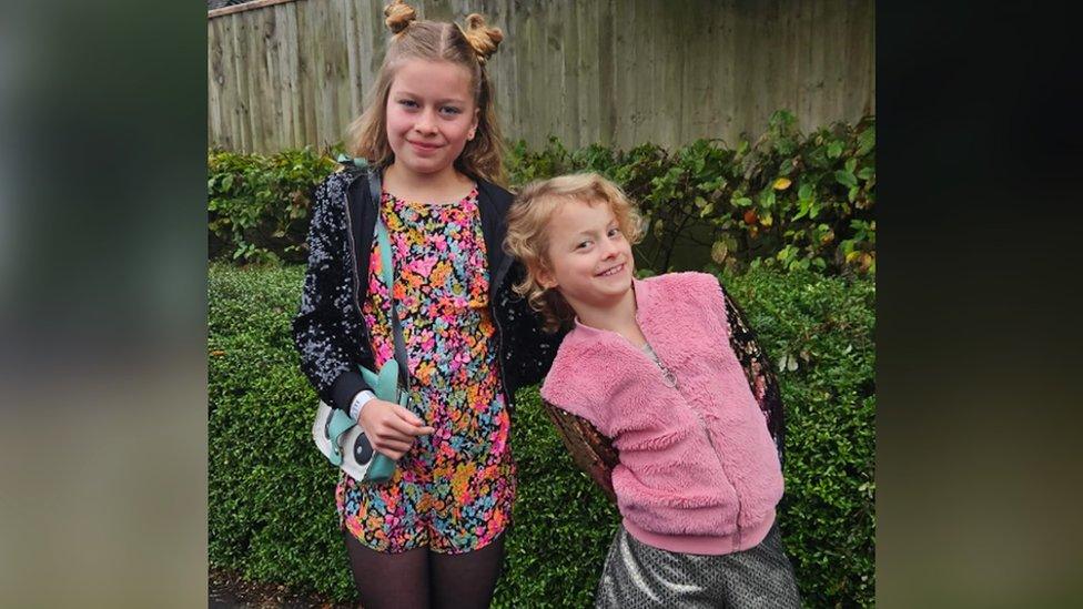 Image of Thea and Lila. They are stood outside together, looking directly at the camera and smiling. Lila is leaning sideways, on Thea's arm.