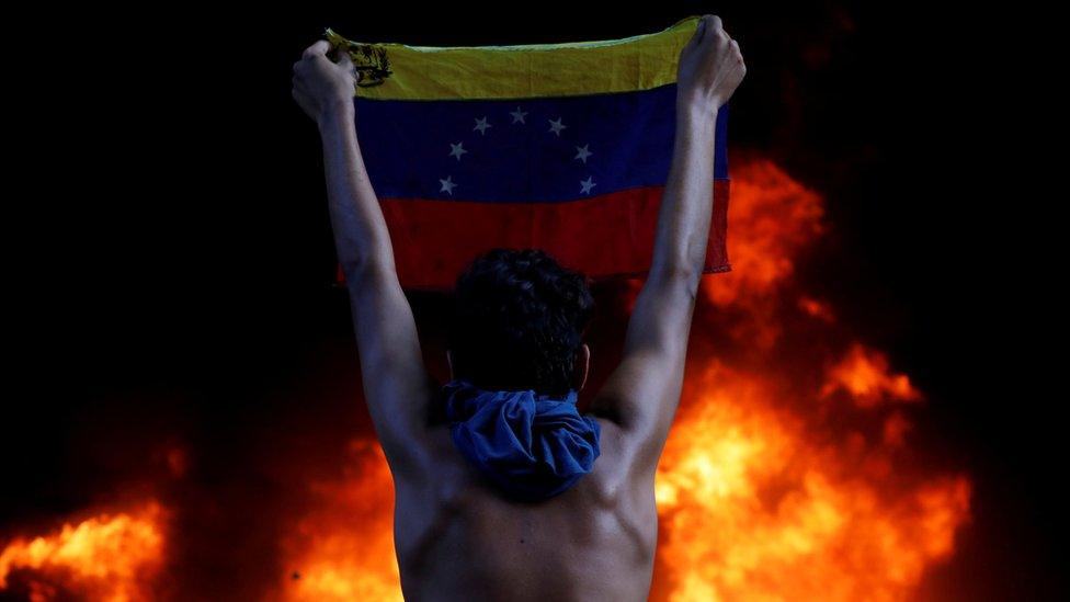 A protestor holds a national flag while standing in front of a fire burning at the entrance of a building housing the magistracy of the Supreme Court of Justice and a bank branch.