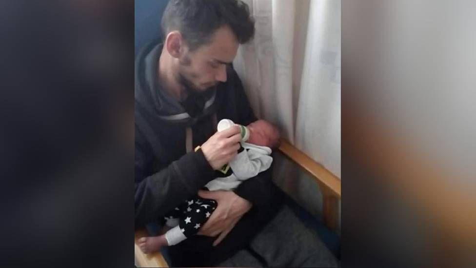 A man with brown hair holding a baby in his left arm. With his right hand, he is feeding the child with a bottle of milk. They are sitting in a chair with a curtain visible on the left of the pair of them.