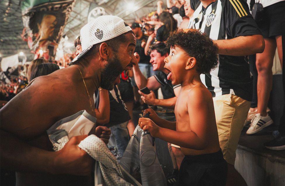 A father and child celebrate Atletico Mineiro scoring. They are both screaming at each other with joy. They are surrounded by cheering supporters in black-and-white striped jerseys 
