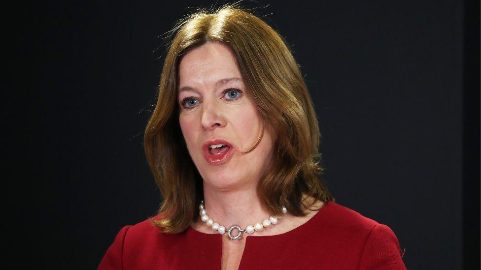 Dr Catherine Calderwood, with brown hair and wearing a red top and white pearl necklace, speaks in front of a dark background at a press conference 