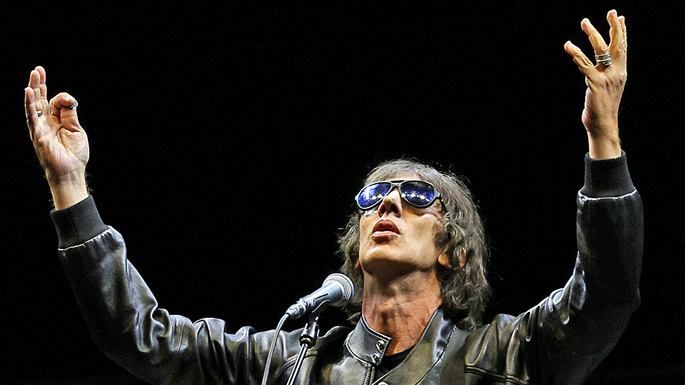 Richard Ashcroft in black jacket and sun glasses holds his hands aloft and looks skywards while taking to the stage