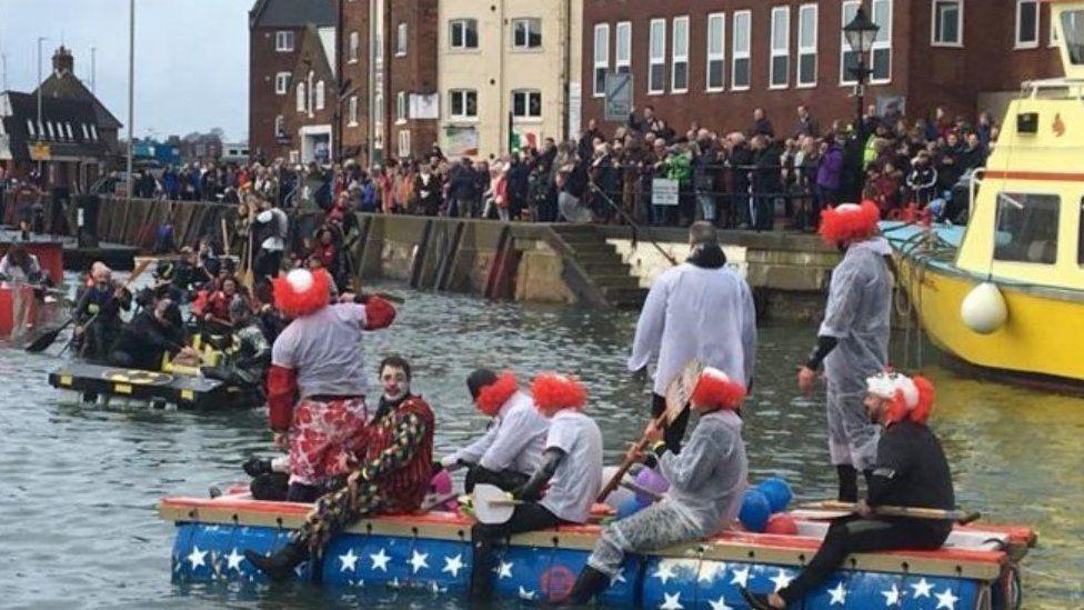 Crowds along a sea wall with people in fancy dress on the water aboard raft-type boats and bright yellow large boat to the right.