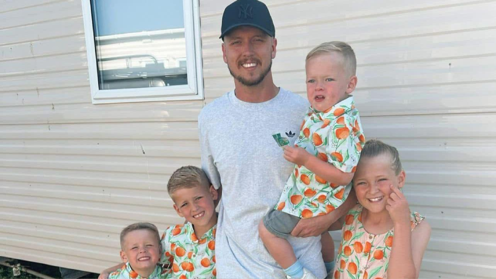 Nathan smiling at the camera. He is holding one of his young sons with one arm, with his daughter to one side of him and his two other sons on the other side. He is wearing a dark baseball cap with the New York Yankees logo on and a grey t-shirt. His sons are all wearing matching button-up shirts with oranges on, while his daughter has a dress made of the same patterned material. They are standing outside what looks like a cream-coloured corrugated caravan wall with a window in the top left corner. 