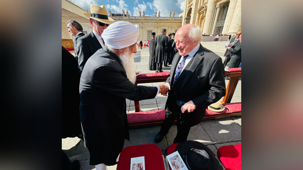 A bearded man, Professor Mohinder Singh, is shaking hands with a man, Irish President Michael D Higgin, who has white hair and is wearing a dark suit with a white shirt and a colourful tie.
