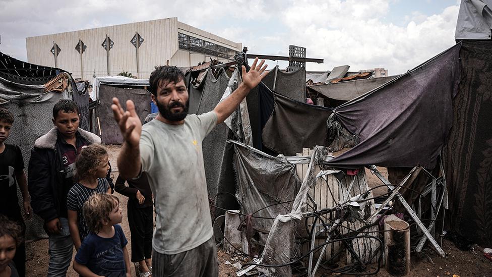 A man in a dirty looking grey T-shirt and trousers with dark hair and a beard standing with his arms outstretched in front of a makeshift shelter of metal strips and large sheets of dark cloth. Behind him stand a group of children.