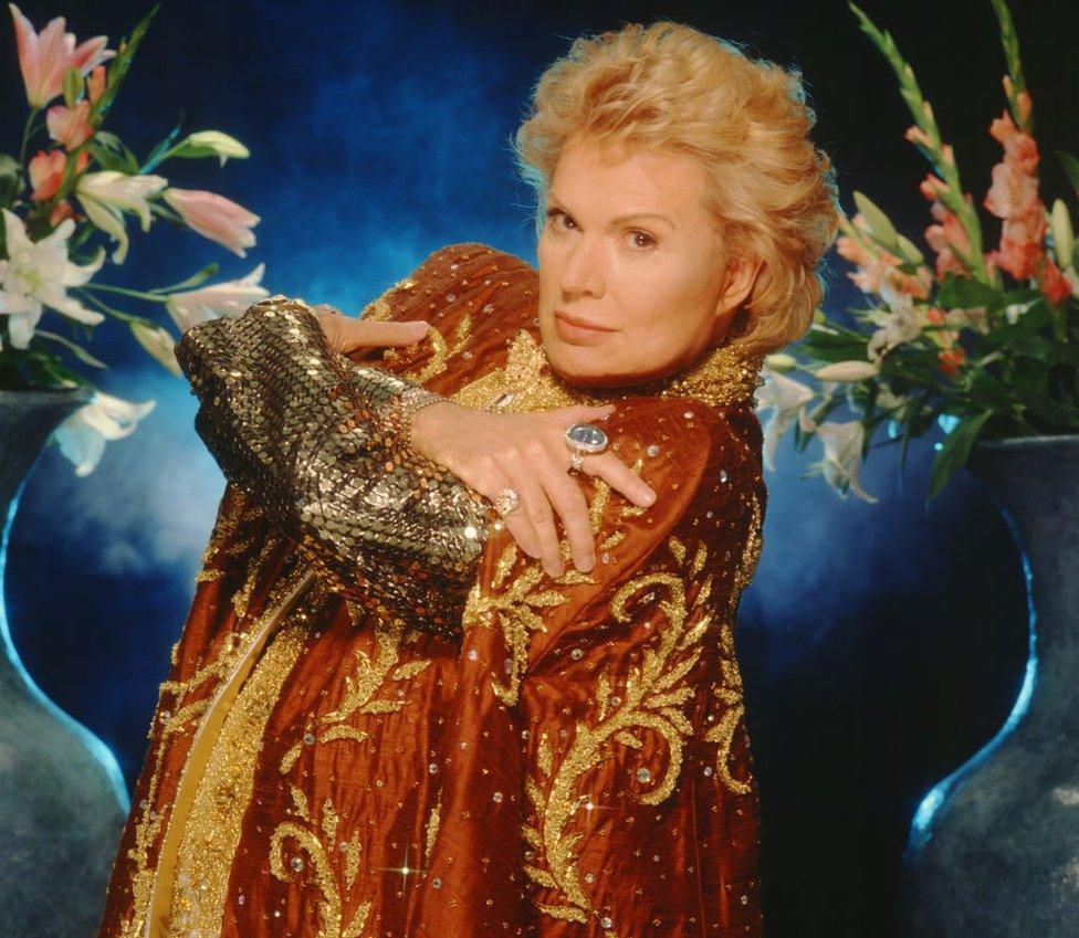 Walter Mercado poses with his hands crossed over his chest, in a sequinned orange and gold robe