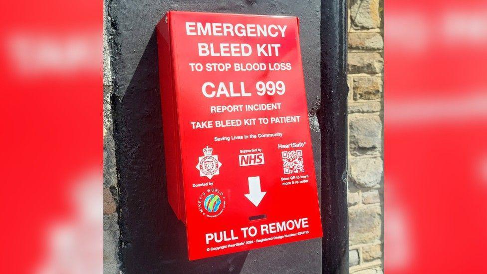 A bleed kit on a wall - it is red and has a lot of wording on it, including the sponsors and "emergency bleed kit to stop blood loss - call 999 - report incident - take bleed kit to patient