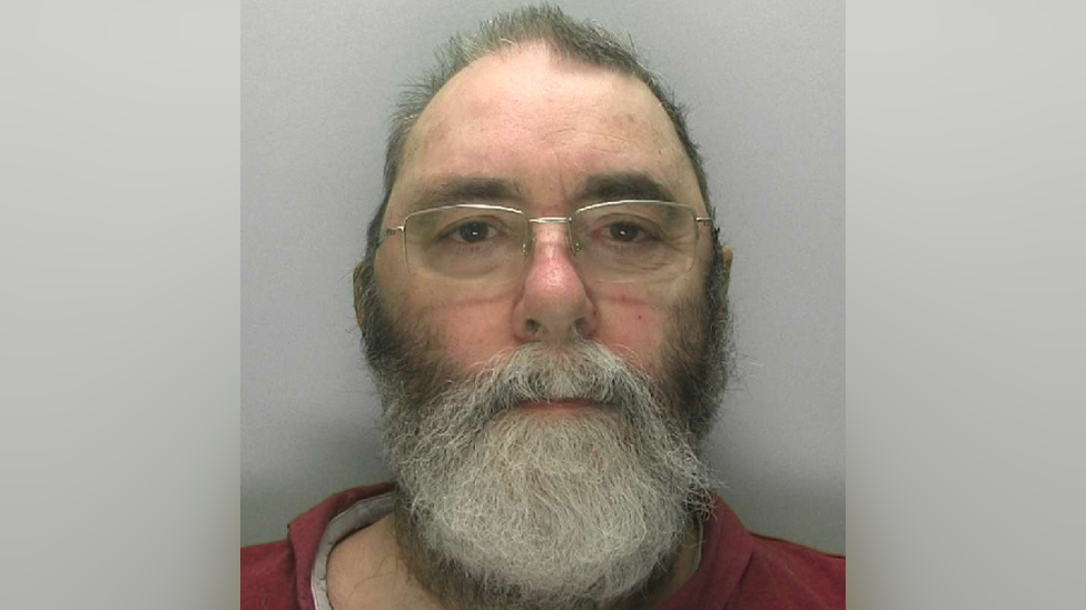 Mugshot of David James. He has grey short hair and a long grey beard. He is wearing glasses and looking directly at the camera with a blank expression