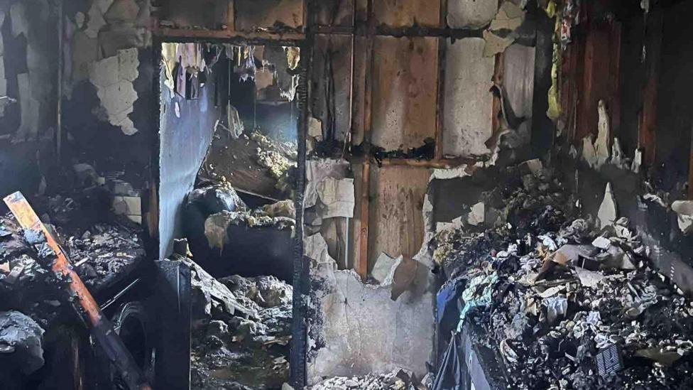 A kitchen which has been gutted and has severe fire damage. The walls have been destroyed. A door frame looks into the living where you can see the sofa has been completely destroyed by a fire. 