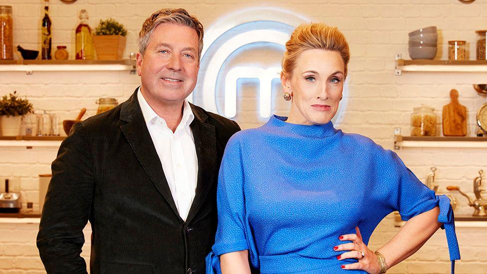 John Torode and Grace Dent stand side by side in a kitchen setting in a publicity shot for Celebrity Masterchef. John is wearing a black jacket over a white shirt, while Grace is dressed in a blue outfit with short sleeves and tied cuffs. Behind them, there are shelves with various kitchen items and a large illuminated logo on the wall.