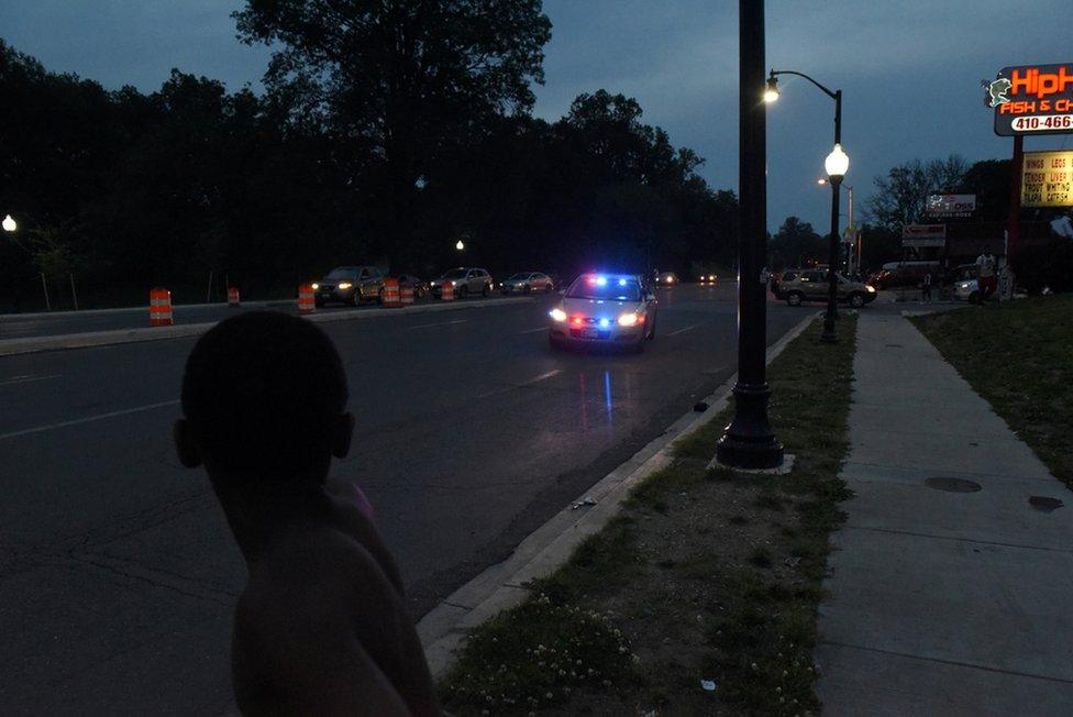 A young boy watches as a police car approaches while they gather in the parking lot of Hip Hop Fish and Chicken in Baltimore