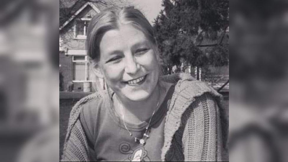 A black and white picture of Dawn Sturgess smiling at the camera, wearing a t-shirt, cardigan and a necklace. She is sitting on the grass in front of a house and a tree.