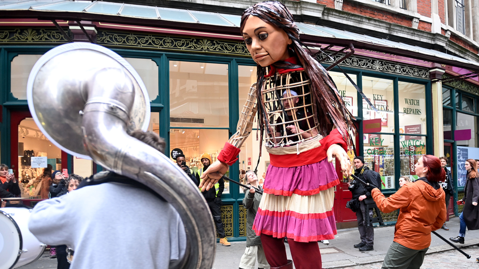 Burning off her lunch, Little Amal has a dance with her friends from Das Brass brass band