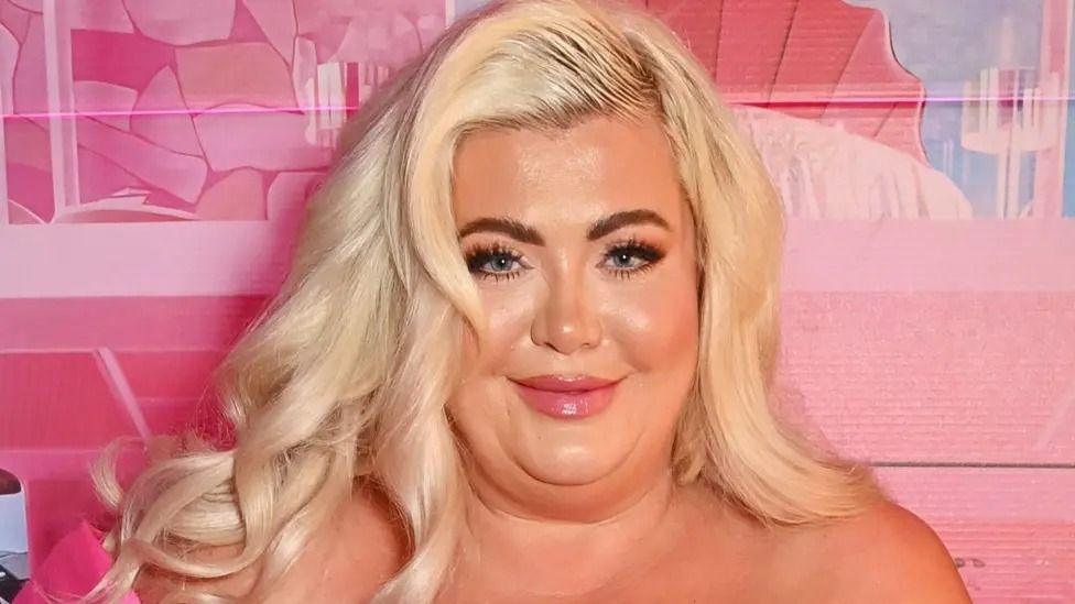 Gemma Collins is photographed looking into the camera at an event. She has dark brown eyes, long eyelashes and long, bleached blonde hair. The background wall behind her is different shades of pink.