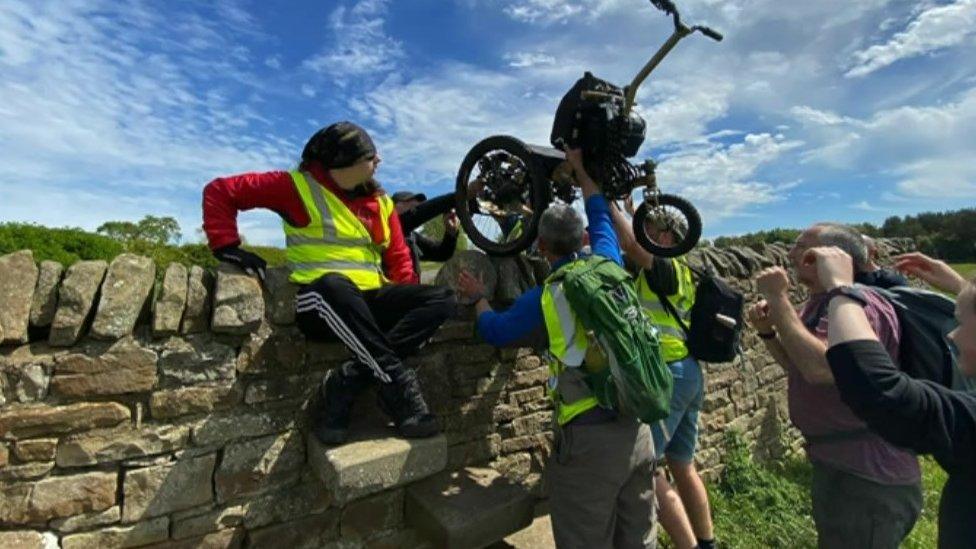 Brynn's wheelchair is lifted over the wall by his support team