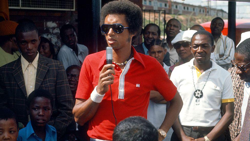 Arthur Ashe, wearing a red shirt, beaded necklace and dark sunglasses, holds a microphone as he talks to South Africans who crowd around him on a visit to Soweto where he ran a tennis clinic - November 1973.