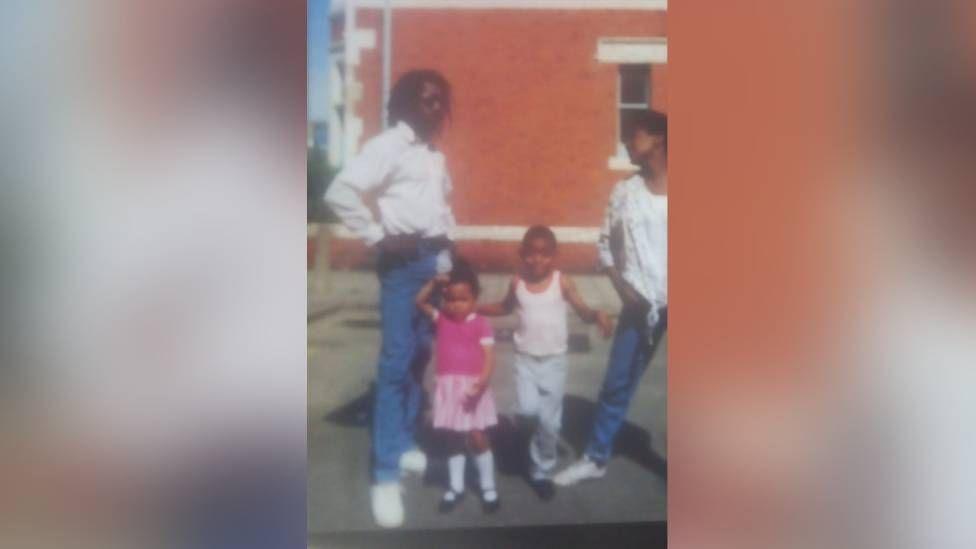 A group shot of two adults standing in a street with two children between them. Both adults are wearing white shirts and jeans, while the little girl is wearing a pink dress with long white socks, and the boy is wearing a white vest and jeans.