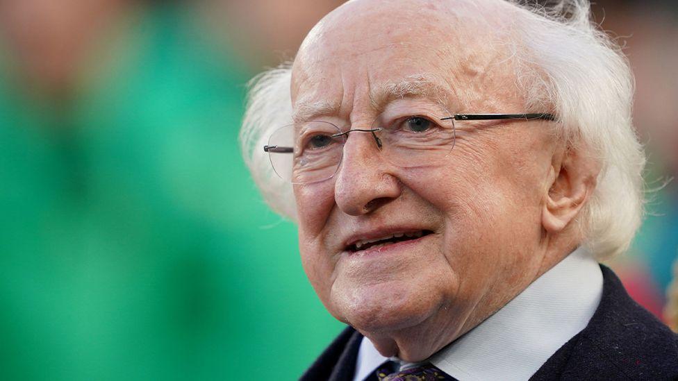 Michael D Higgins standing looking to camera at an event in Dublin. He is dressed in a dark coloured suit with grey shirt and floral tie.