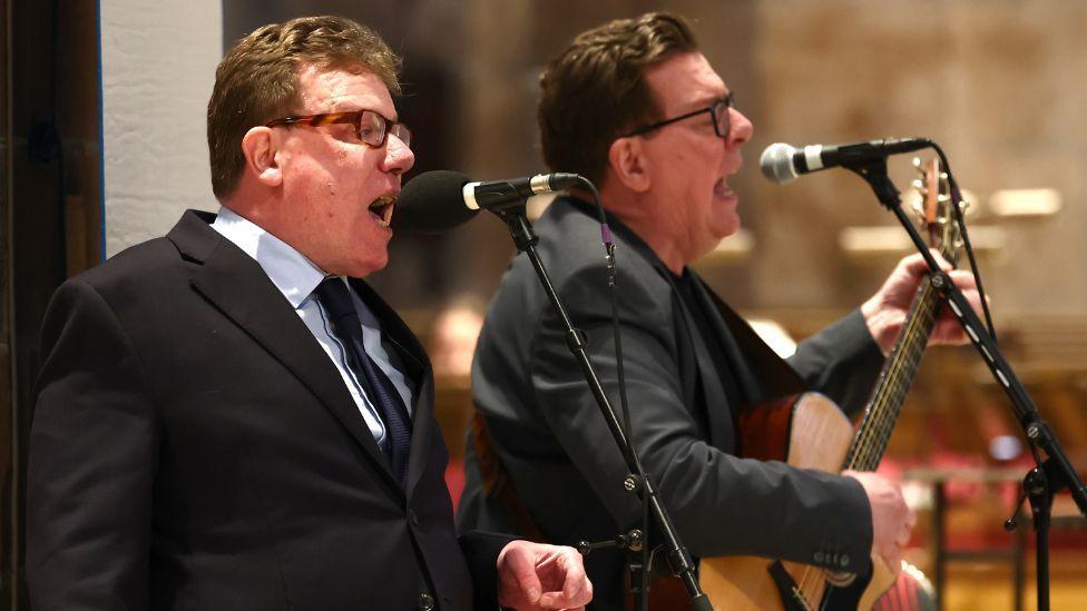 The Proclaimers, twins with dark hair, dark glasses and dark suits, sing into microphones alongside each other on stage. 