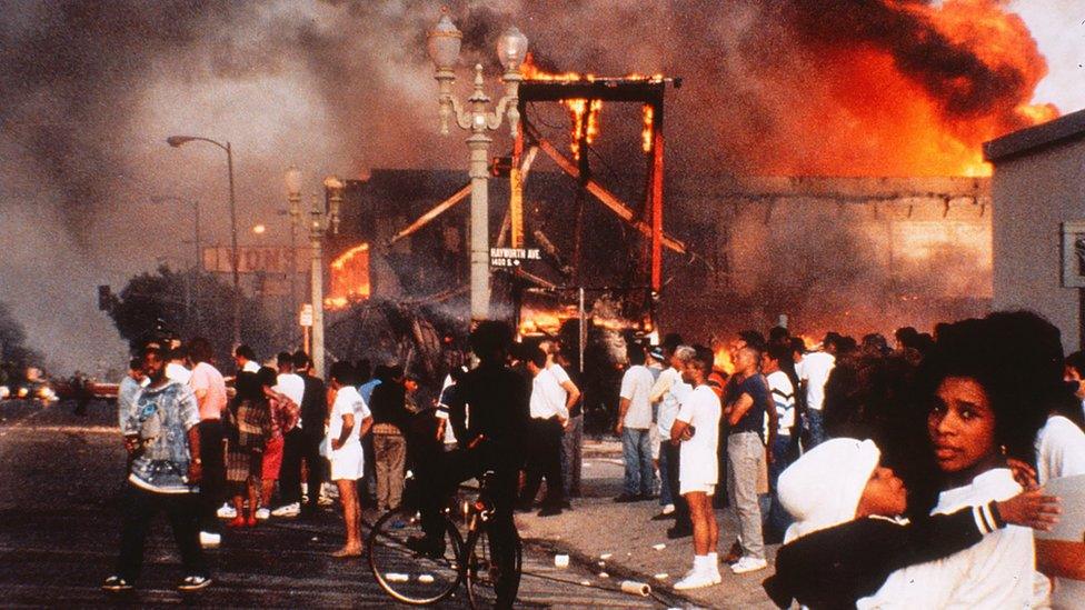 Mother and child amidst mayhem, businesses burning, bystanders watching raging fires, pedestrian walking in the street drinking from a 40-ounce at intersection of Pico Boulevard and Hayworth Avenue in West Hollywood, the sky black with smoke in daylight on April 30, 1992