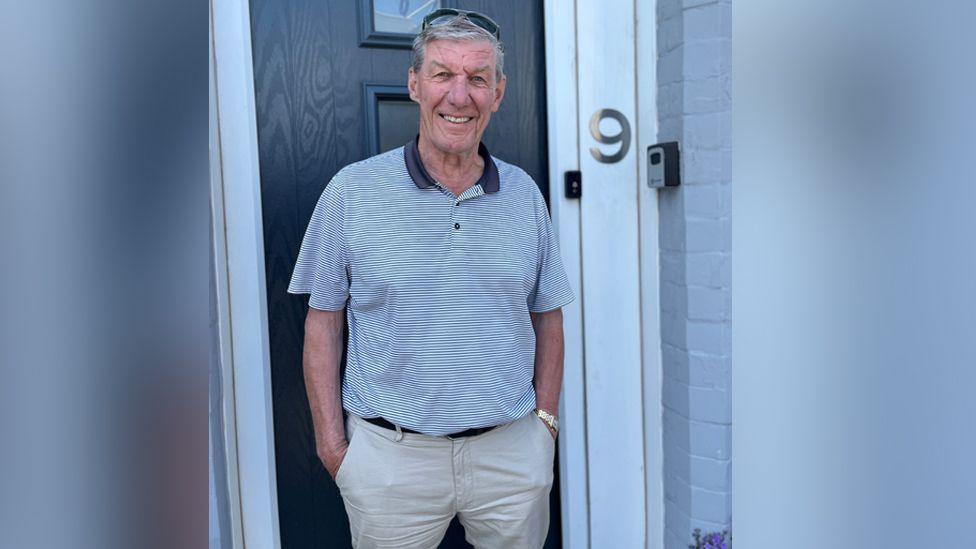 Ray is standing in the doorway of Goodison Road and is smiling directly at the camera. The bricks of the property are painted in white and the door is black.