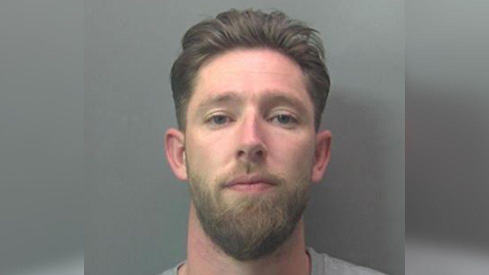 A police mugshot of Matthew Fox. He has a beard and swept-back hair and is looking straight at the camera. He is wearing a grey top and is in front of a grey background.